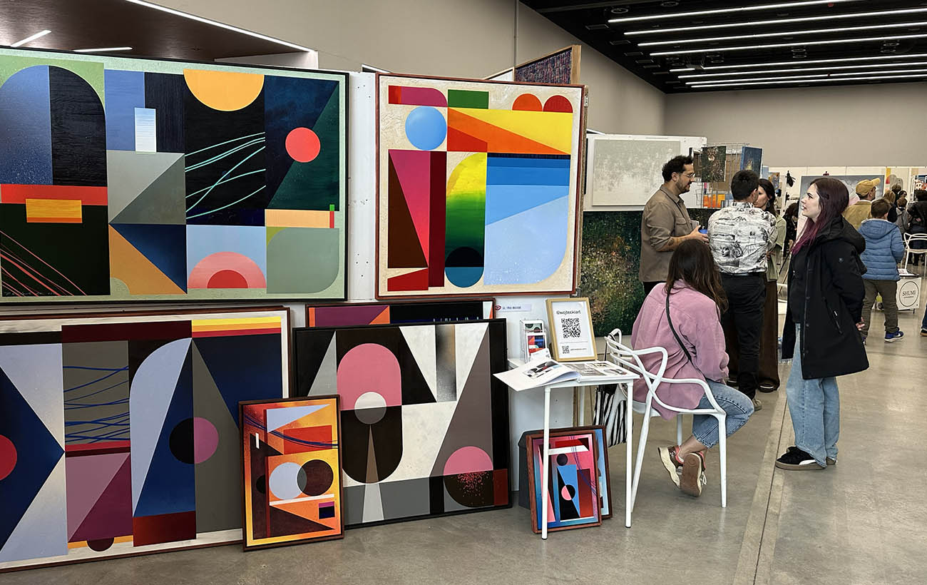 Perspektywa tętniących życiem targów sztuki. Przestronne, minimalistyczne stoiska z bogatą ekspozycją malarstwa, tworzące atmosferę kulturalnego wydarzenia i dialogu ze sztuką.